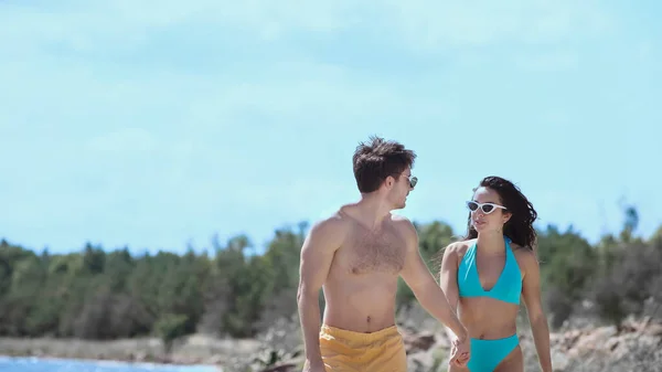 Young man in sunglasses holding hand of girlfriend in swimsuit on beach — Stock Photo