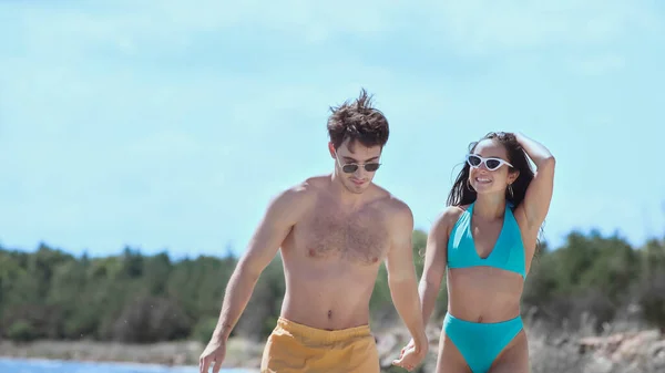 Позитивная молодая пара в купальниках, держащаяся за руки на пляже — стоковое фото