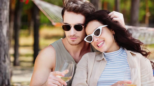 Hombre en gafas de sol tocando el pelo de novia sonriente con vino en la playa - foto de stock