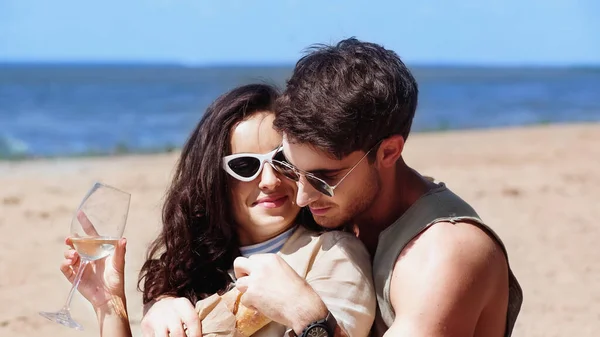 Young man hugging girlfriend in sunglasses with wine and baguette on beach — Stock Photo