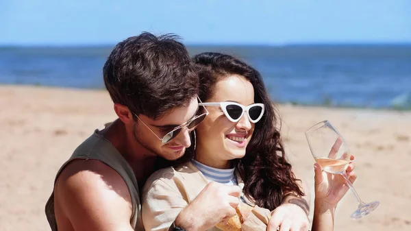 Cheerful woman in sunglasses holding wine near boyfriend with baguette on beach — Stock Photo