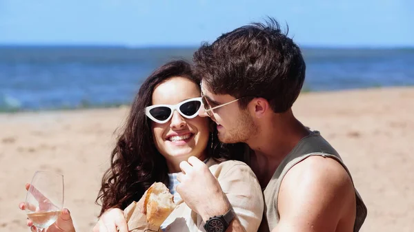 Smiling man in sunglasses hugging girlfriend with wine and baguette on beach — Stock Photo