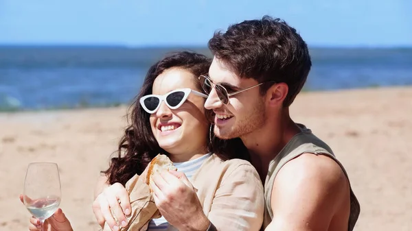 Cheerful man in sunglasses holding baguette and hugging girlfriend with wine on beach — Stock Photo