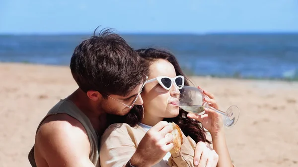 Frau mit Sonnenbrille trinkt Wein neben Freund mit Baguette am Strand — Stockfoto