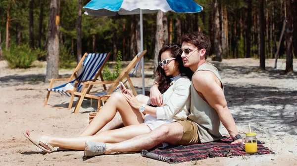 Uomo in occhiali da sole abbracciare fidanzata con smartphone vicino succo d'arancia sulla spiaggia — Foto stock