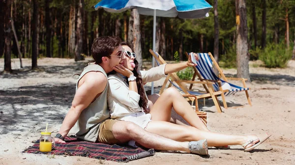 Pareja positiva en gafas de sol tomando selfie en manta en la playa - foto de stock