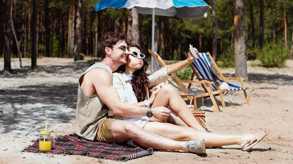 Улыбающийся мужчина в солнечных очках делает селфи рядом с девушкой на одеяле на пляже — стоковое фото