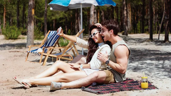 Щасливий чоловік в сонцезахисних окулярах обіймає подругу, приймаючи селфі на пляжі — стокове фото