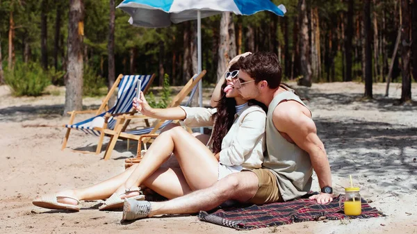 Woman in sunglasses taking selfie on smartphone and sticking out tongue near boyfriend on beach — Stock Photo