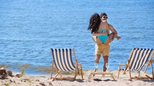 Hombre sonriente en gafas de sol abrazando a su novia en traje de baño en la playa - foto de stock