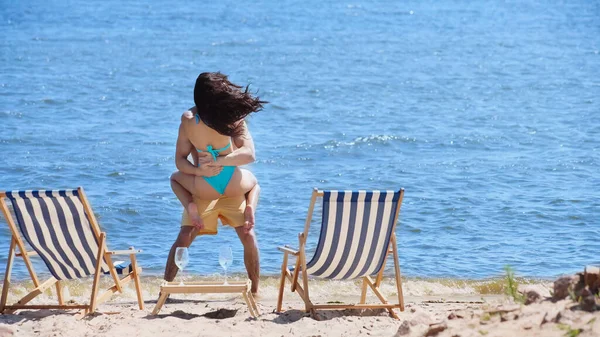 Man holding girlfriend in swimsuit near deck chairs on beach — Stock Photo