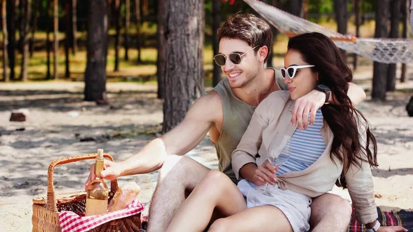Улыбающийся мужчина обнимает девушку в солнечных очках и берет бутылку вина на пляже — стоковое фото