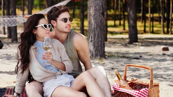 Junge Frau mit Sonnenbrille hält Wein neben Freund und Korb am Strand — Stockfoto