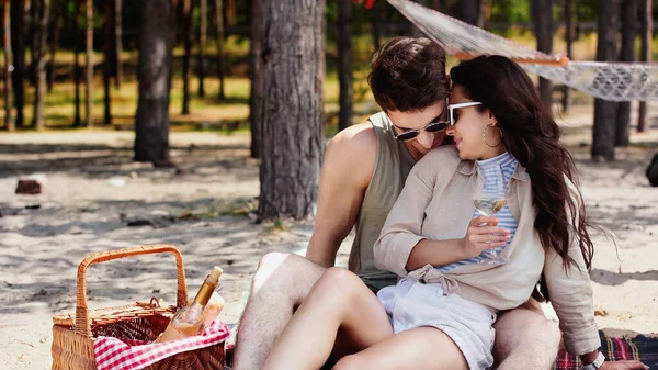 Mujer joven en gafas de sol sosteniendo el vino y sentado cerca de novio en manta en la playa - foto de stock