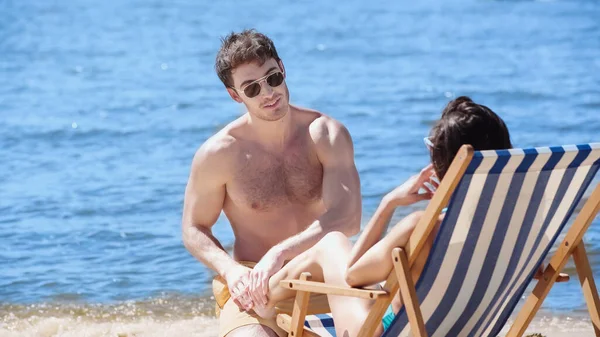 Smiling man in sunglasses touching leg of girlfriend on deck chair on beach — Stock Photo