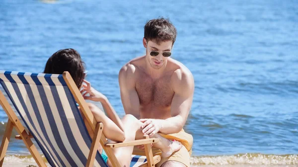 Homme en lunettes de soleil toucher jambe de petite amie sur chaise longue près de la mer sur la plage — Photo de stock