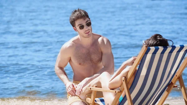 Мужчина в солнечных очках трогает ногу подруги на шезлонге на пляже возле моря — стоковое фото