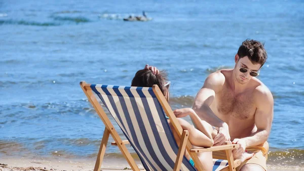 Man in swimwear touching leg of girlfriend on deck chair on beach — Stock Photo