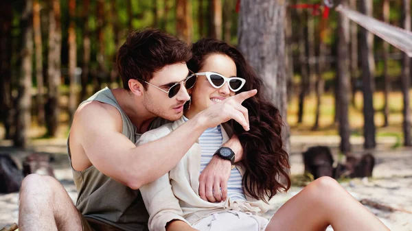 Mann mit Sonnenbrille umarmt Freundin und zeigt mit Finger auf Strand — Stockfoto