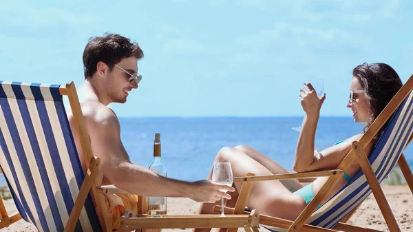 Улыбающаяся женщина в купальнике держит бокал вина рядом с парнем на шезлонге на пляже — стоковое фото