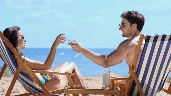 Smiling couple in sunglasses clinking wine on deck chairs on beach — Stock Photo