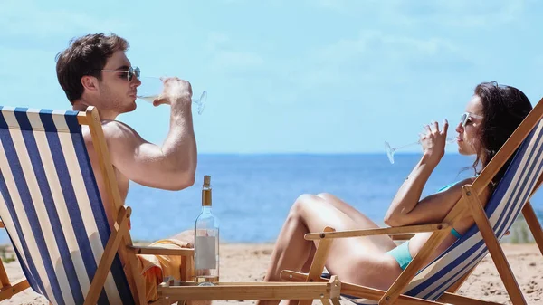 Pareja en gafas de sol bebiendo vino en tumbonas en la playa - foto de stock
