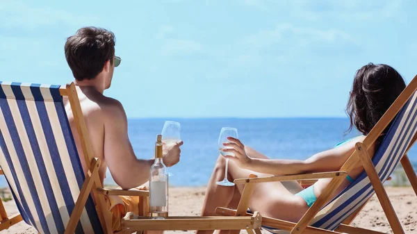 Pareja joven sosteniendo copas de vino en tumbonas en la playa - foto de stock