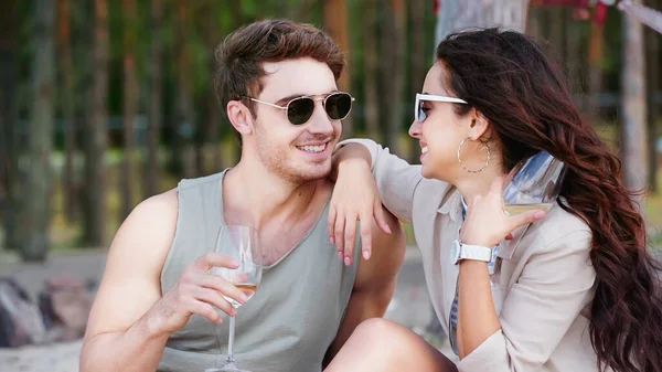 Mulher sorridente em óculos de sol olhando para o namorado com vinho na praia — Fotografia de Stock