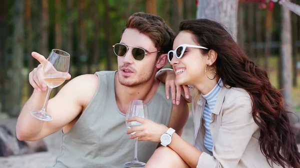 Giovane uomo che tiene un bicchiere di vino e punta con il dito vicino alla ragazza in occhiali da sole sulla spiaggia — Foto stock