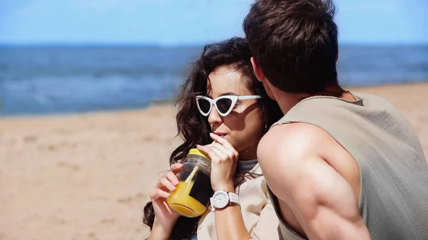 Brunette woman in sunglasses drinking orange juice near boyfriend on beach — Stock Photo