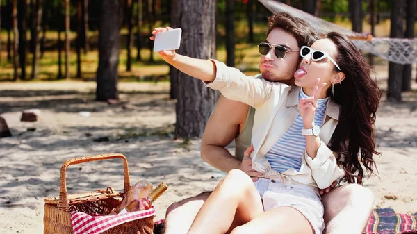 Pareja joven tomando selfie en el teléfono inteligente cerca de la cesta de picnic en la playa - foto de stock