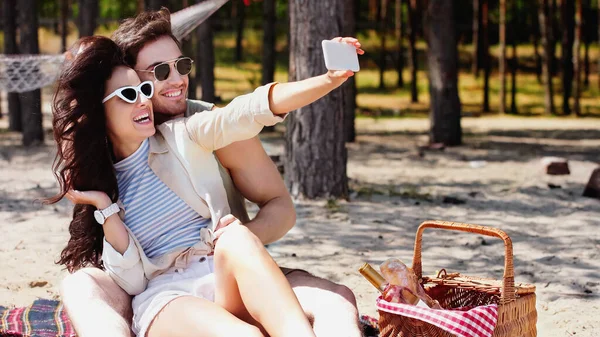 Pareja feliz en gafas de sol tomando selfie en smartphone cerca de la cesta en la playa - foto de stock