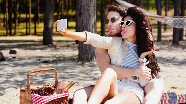 Pareja en gafas de sol haciendo muecas mientras toma selfie en el teléfono inteligente cerca de la cesta de picnic en la playa - foto de stock