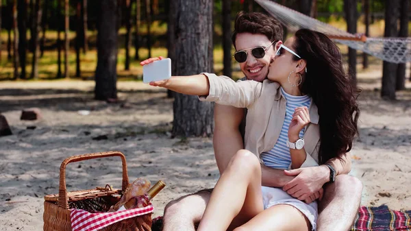 Mujer besando novio en gafas de sol y tomando selfie durante el picnic en la playa - foto de stock