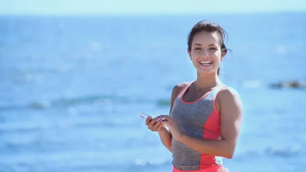 Mujer positiva en ropa deportiva sosteniendo el teléfono celular y mirando a la cámara cerca del mar borroso - foto de stock