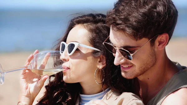 Mulher morena em óculos de sol beber vinho perto de namorado na praia — Fotografia de Stock