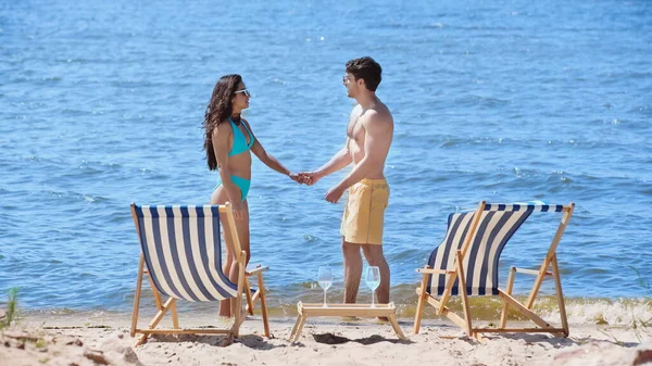 Side view of young couple holding hands near deck chairs and wine on beach — Stock Photo