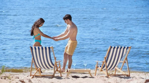 Side view of smiling couple in swimwear holding hands near deck chairs and wine on beach — Stock Photo