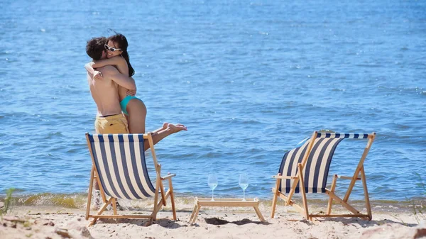 Happy woman in swimsuit hugging boyfriend near wine and deck chairs on beach — Stock Photo