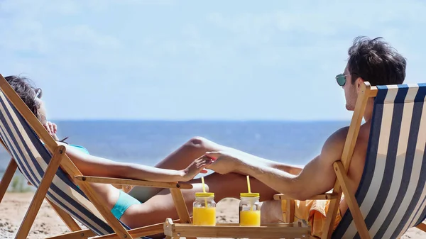 Pareja cogida de la mano en tumbonas cerca de zumo de naranja en frascos en la playa - foto de stock