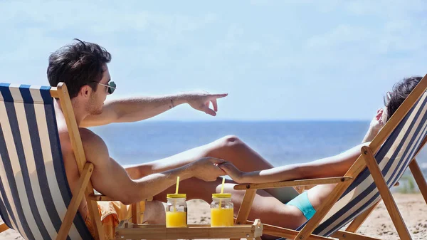 Man in sunglasses holding hand of girlfriend and pointing with finger near orange juice on beach — Stock Photo