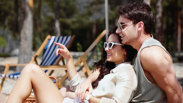 Smiling man embracing girlfriend pointing with finger on beach — Stock Photo