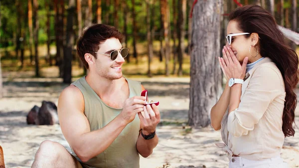 Улыбающийся мужчина, держащий ювелирное кольцо рядом с девушкой в солнечных очках на пляже — стоковое фото