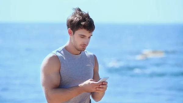 Athletic sportsman using smartphone near blurred sea — Stock Photo