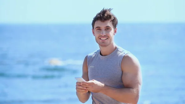 Deportista positivo usando teléfono inteligente y mirando a la cámara cerca del mar borroso - foto de stock