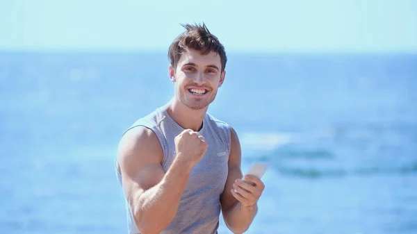 Esportista alegre mostrando sim gesto e segurando smartphone perto do mar borrado — Fotografia de Stock