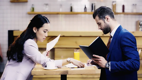 Asian businesswoman looking at documents near bearded businessman in cafe — Stock Photo