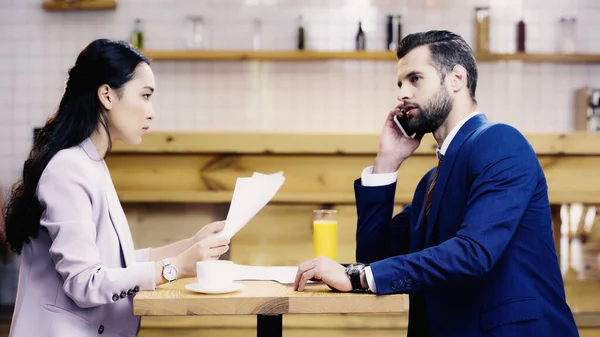 Азиатская бизнесвумен держит документы рядом с бизнесменом, разговаривающим на смартфоне в кафе — стоковое фото