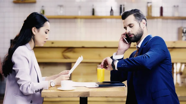 Mujer de negocios asiática mirando documentos cerca de hombre de negocios hablando en el teléfono inteligente y comprobar el tiempo en la cafetería - foto de stock