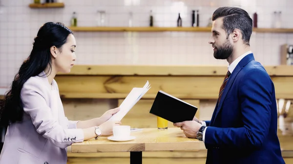 Vista lateral da mulher de negócios asiática segurando documentos perto de empresário no café — Fotografia de Stock